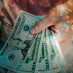 Mão segurando notas de dólar com um efeito de luz dourada e brilho ao fundo, simbolizando abundância e conexão energética com o dinheiro.
